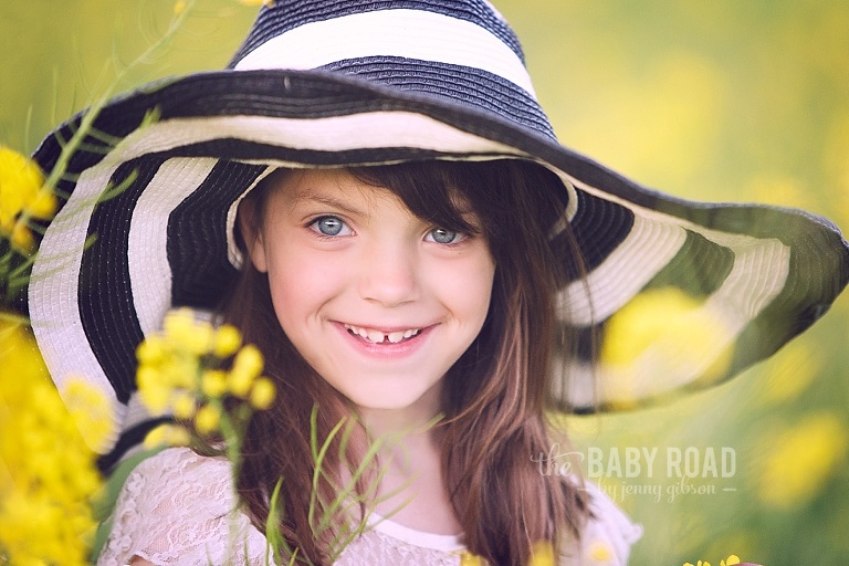 Little girl in black and white striped hat smiling in a meadow of yellow mustard flowers